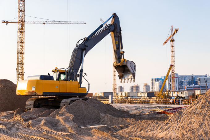 Ein gelber Bagger steht zwischen mehreren Sandhaufen auf einer Baustelle. Im Hintergrund sind Kräne und Wohnblocks zu sehen.